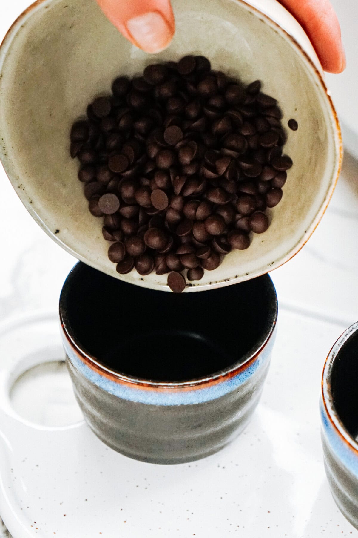 A hand is pouring chocolate chips from a bowl into a blue and black mug, ready to create topping for gourmet caramel apples.