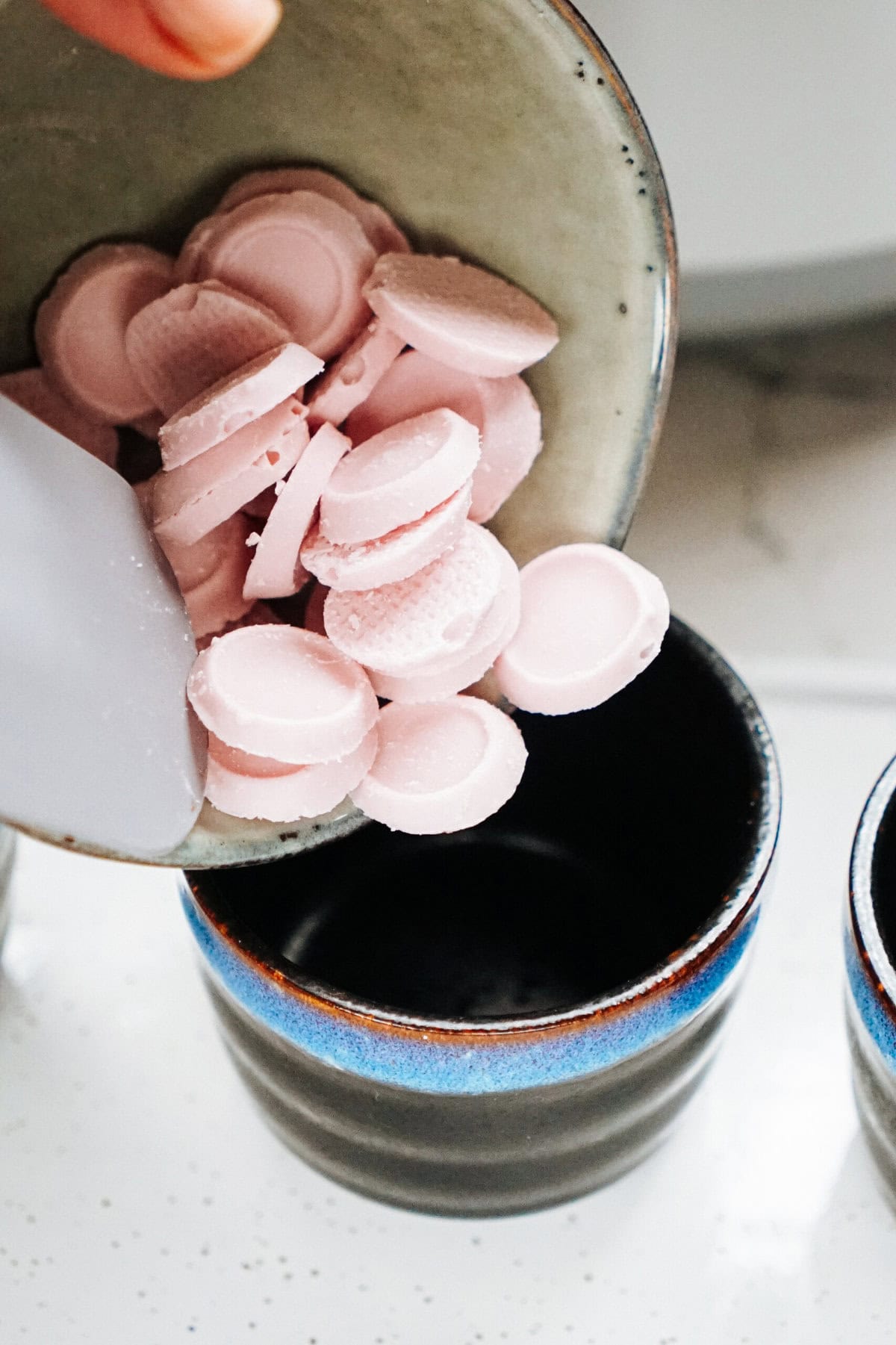 A hand is pouring small, round, pink pieces from a plate into a dark cup on a white countertop, evoking the meticulous craft akin to preparing gourmet caramel apples.