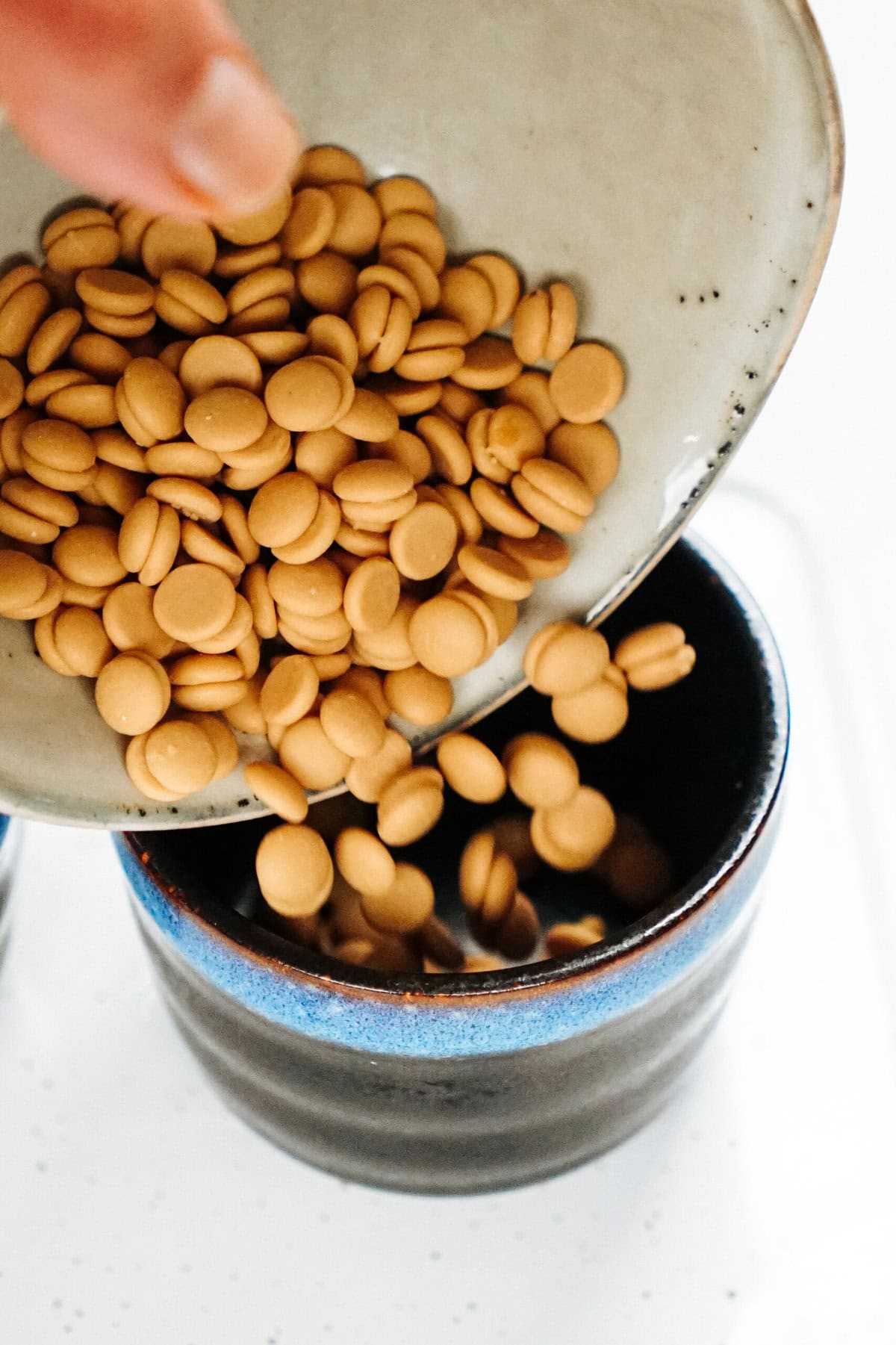 A hand is pouring a bowl of small, round, beige pellets into a black and blue cup, reminiscent of the delicate precision used in crafting gourmet caramel apples.