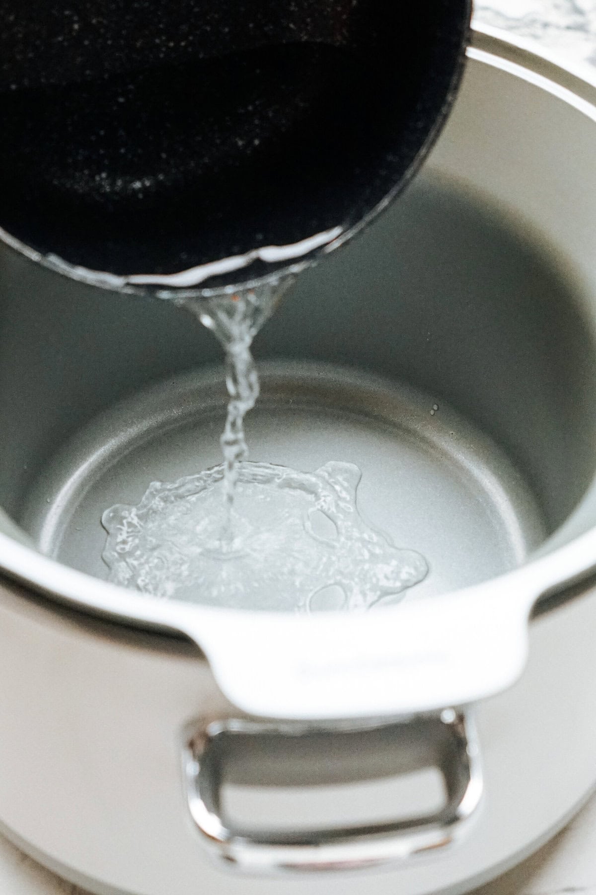 Water being poured from a black pan into a silver pot on a stovetop, preparing the base for making gourmet caramel apples.