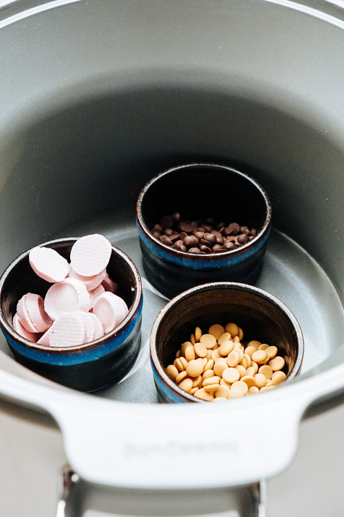 Three small bowls with pink, brown, and yellow chocolate buttons are placed inside a larger pot, likely for melting—perfect for drizzling over gourmet caramel apples.