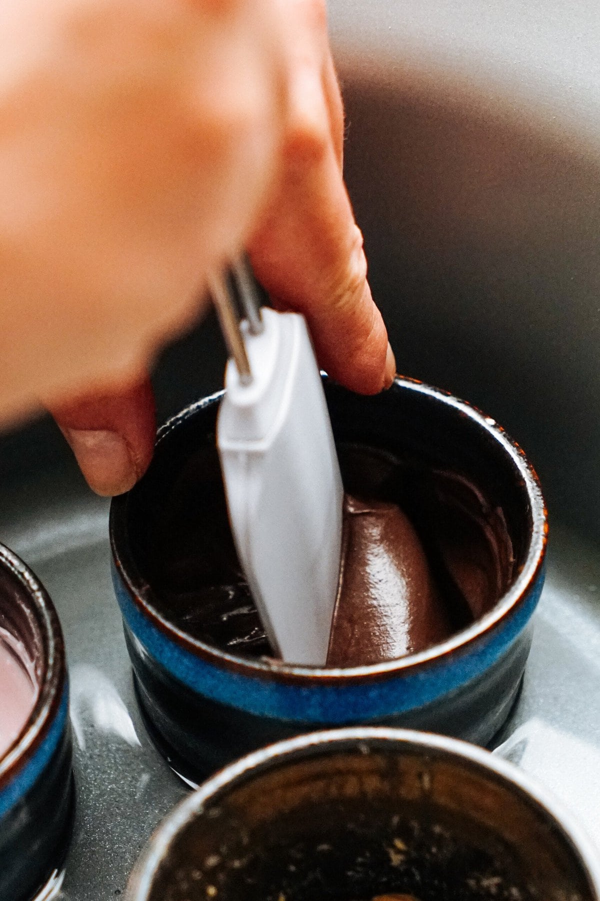 Close-up of a hand whisking dark brown liquid in a small blue ceramic bowl with a handheld mixer, perfect for preparing gourmet caramel apples.