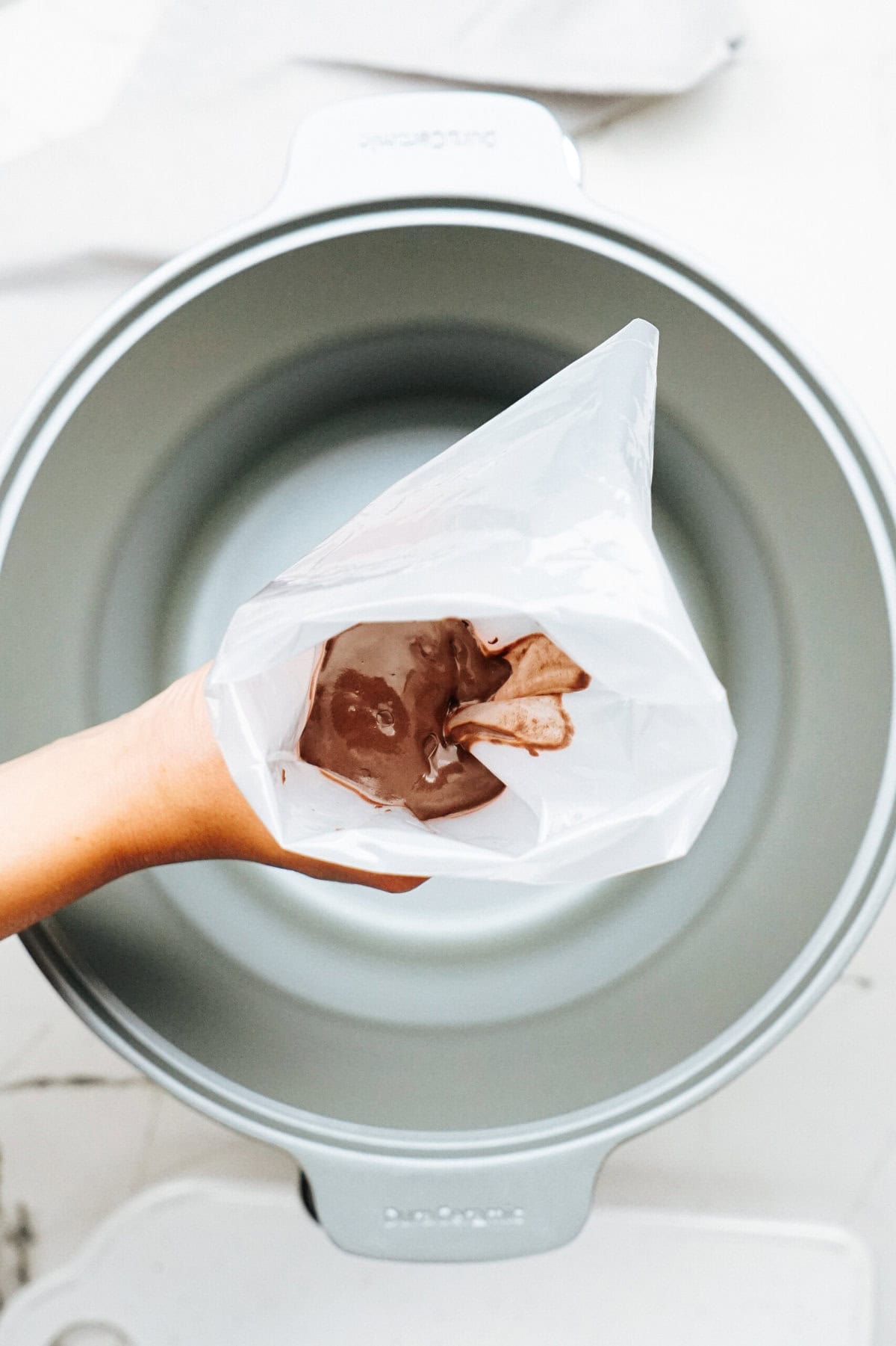 A hand is holding a transparent plastic bag filled with a chocolate substance over a silver mixing bowl, as if preparing to create gourmet caramel apples.