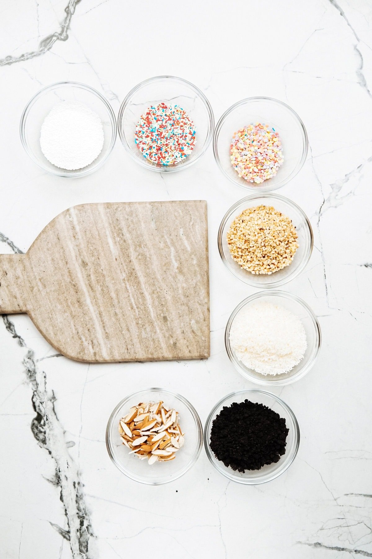 Seven small bowls containing different ingredients arranged around a wooden cutting board: sugar, multicolored sprinkles, white sprinkles, chopped nuts, shredded coconut, sliced almonds, and black sesame seeds—perfect for creating gourmet caramel apples.