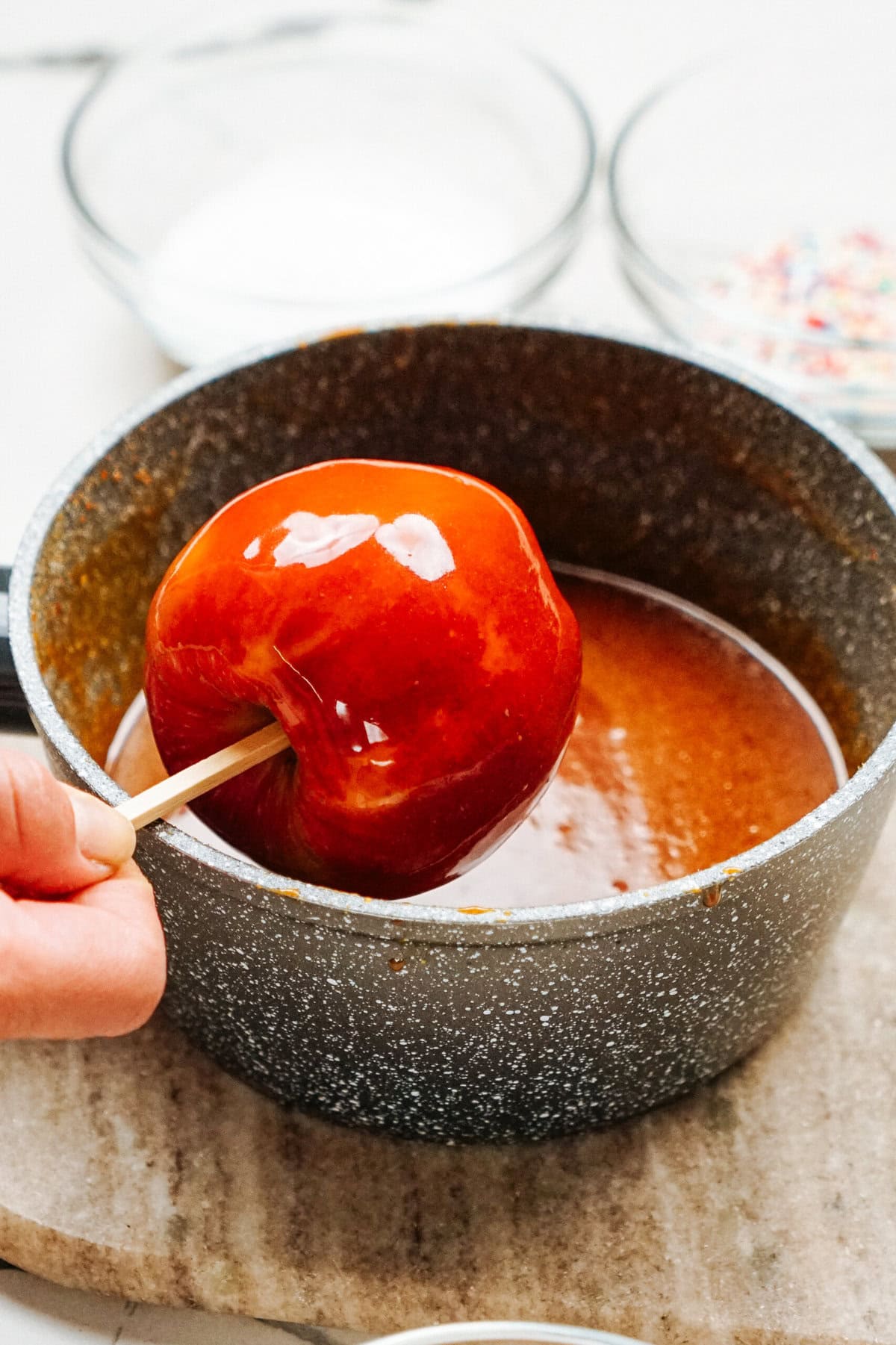 A red candy apple, held by a stick, is being partially submerged in caramel inside a black saucepan. Bowls of sugar and sprinkles sit in the background, all shining with the promise of delicious gourmet caramel apples.