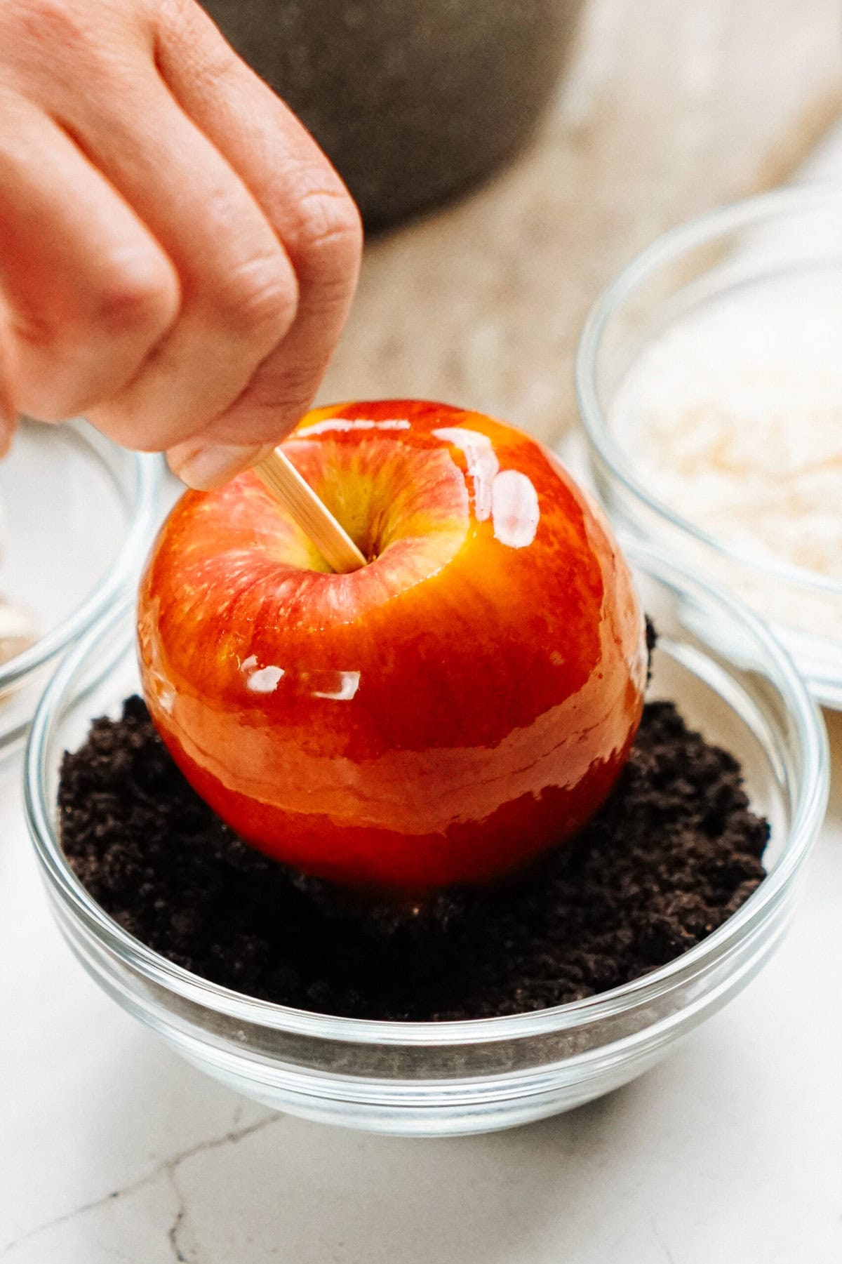 A hand is dipping a shiny red caramel apple into a bowl of crushed cookies, crafting gourmet caramel apples. Two other bowls containing different ingredients are visible in the background.