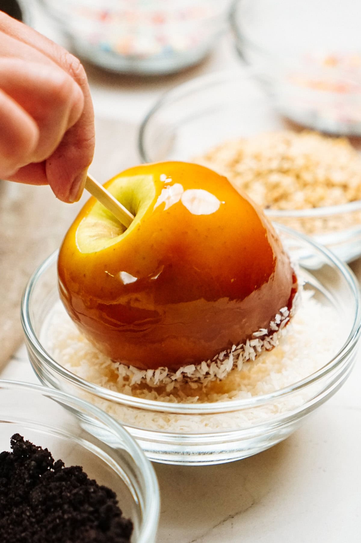 A hand dips a gourmet caramel apple into a bowl of shredded coconut, surrounded by other bowls containing various toppings.