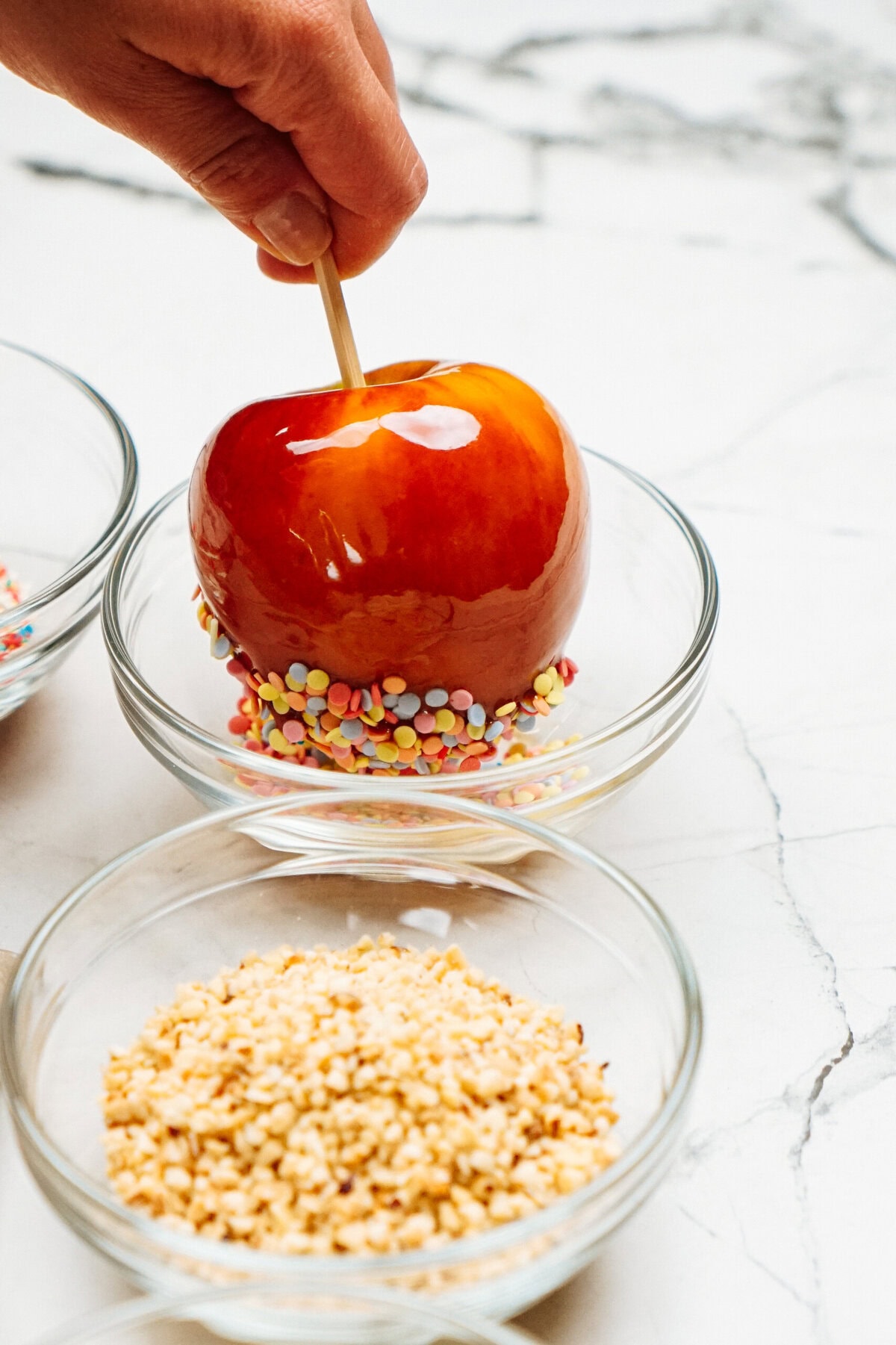 A hand decorates a candy apple with colorful sprinkles by dipping it into a bowl, transforming it into one of the gourmet caramel apples. Another bowl with chopped nuts is nearby.