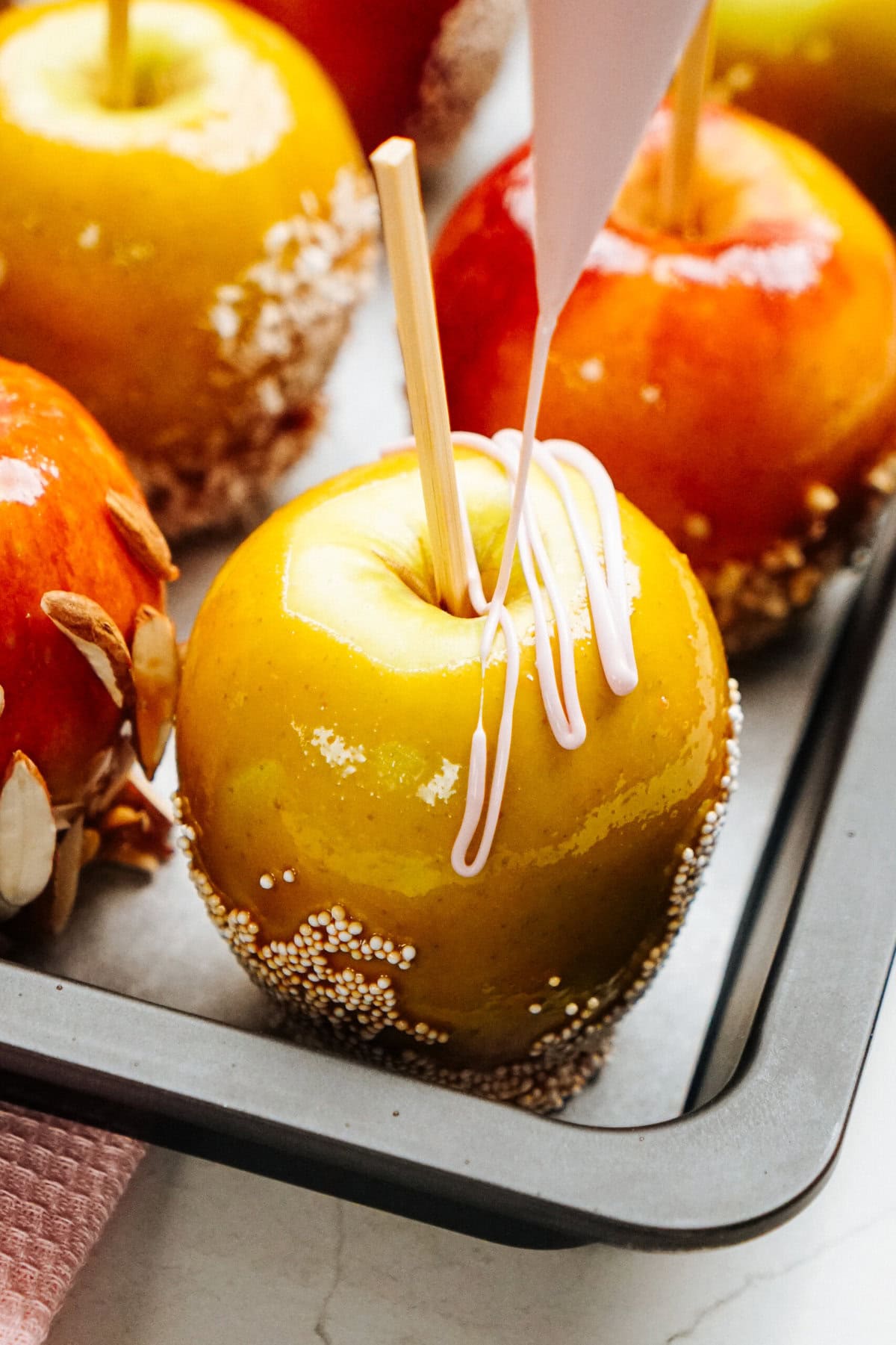 A tray of gourmet caramel apples, some with nut coatings, is being drizzled with white icing from a piping bag.