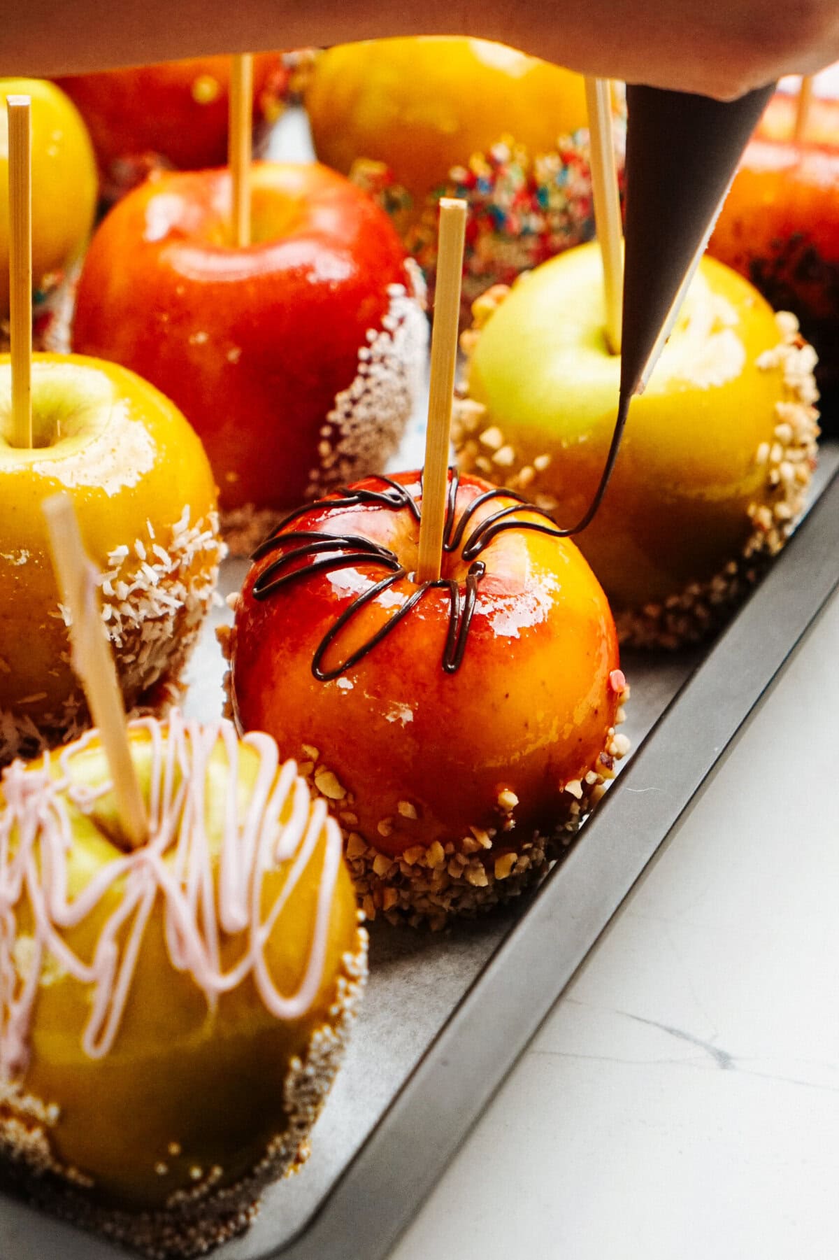 A hand is decorating gourmet caramel apples with chocolate. The apples, mounted on sticks, are coated with various toppings like nuts and sprinkles, and are arranged neatly on a tray.