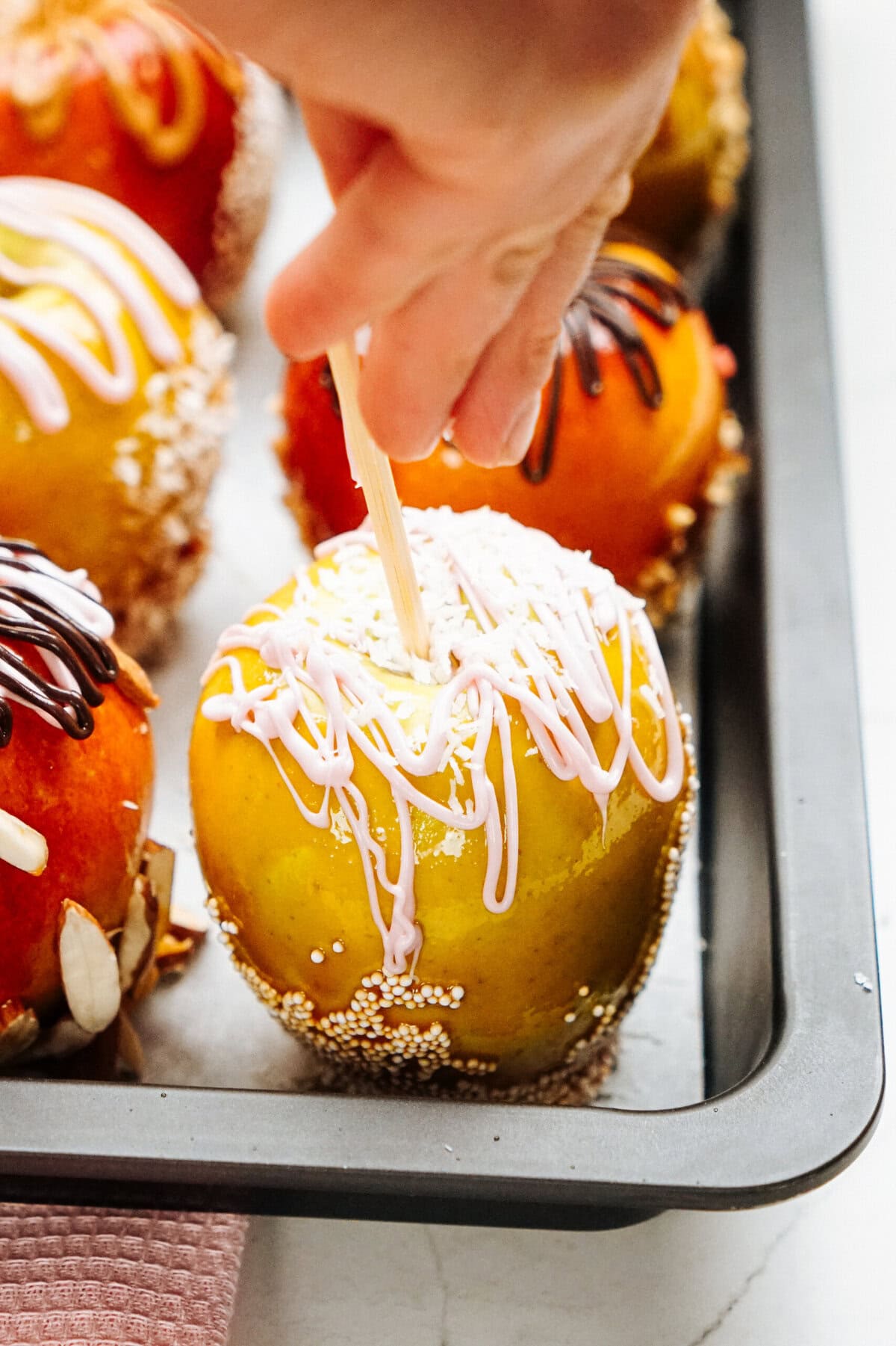 A hand holding a stick is being inserted into a gourmet caramel apple decorated with white drizzle and sprinkles, placed on a baking sheet alongside other similarly adorned apples.