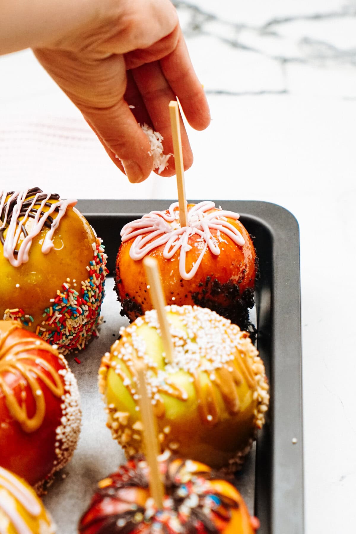 A hand sprinkles coconut flakes onto a gourmet caramel candy apple with a stick on a tray, alongside other decorated candy apples.