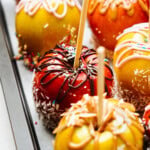 Close-up of multiple gourmet caramel apples on sticks, each decorated with various toppings including sprinkles, chocolate drizzle, and shredded coconut, arranged on a tray.