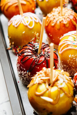 Close-up of multiple gourmet caramel apples on sticks, each decorated with various toppings including sprinkles, chocolate drizzle, and shredded coconut, arranged on a tray.