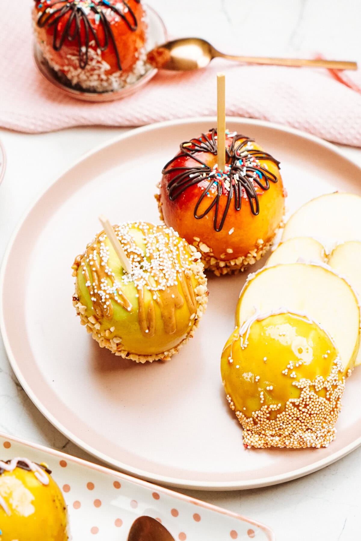 A plate with three gourmet caramel apples, each decorated differently, and slices of a yellow apple. A spoon and napkin are beside the plate.