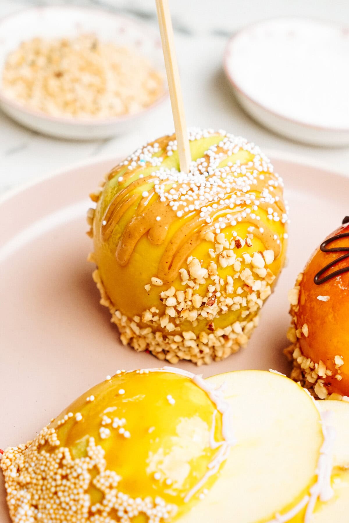 Close-up of a gourmet caramel apple coated with nuts and sprinkles on a plate, with two cut slices of the apple in the foreground and bowls with toppings in the background.