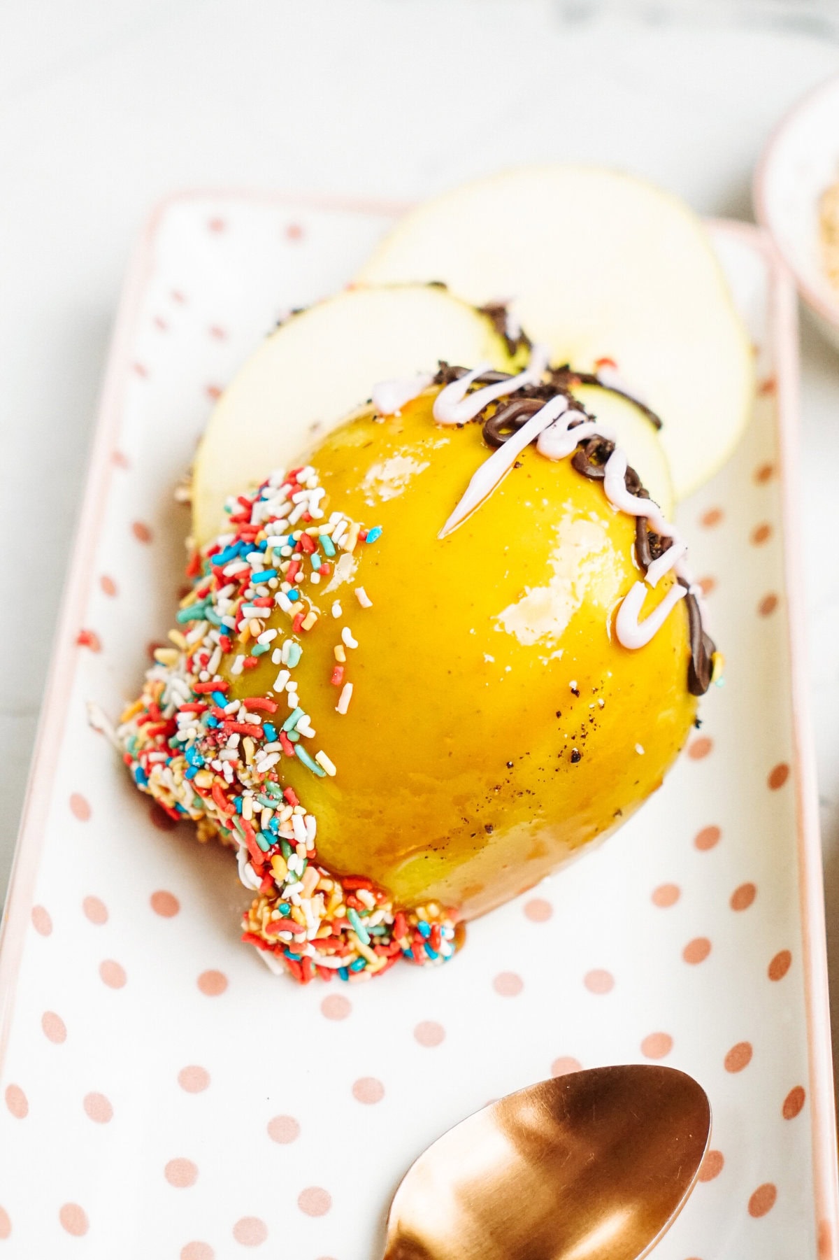 A gourmet caramel apple topped with sprinkles and sliced in half, served on a white rectangular plate with pink polka dots and a gold-colored spoon beside it.