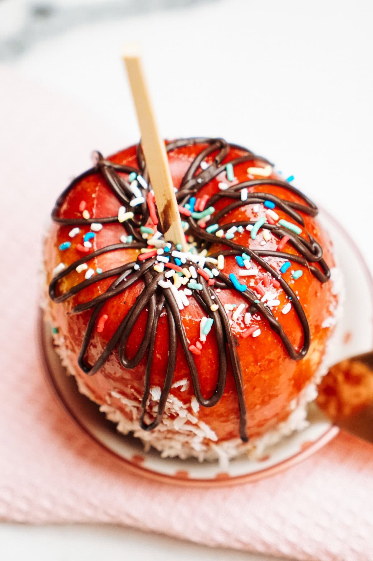 A gourmet caramel apple on a plate, covered with chocolate drizzle and colorful sprinkles, resting on a pink surface.