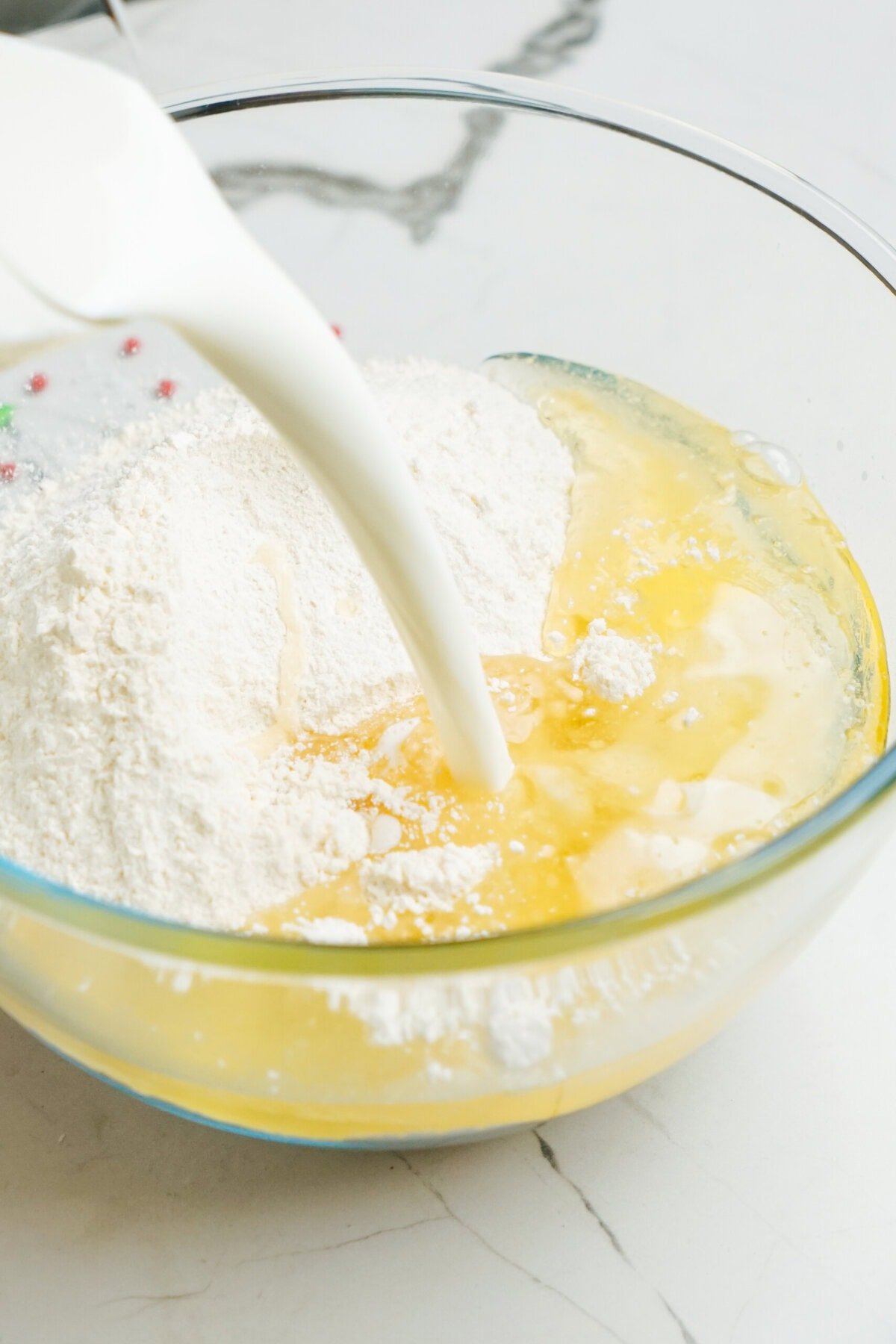 A clear glass bowl with flour and egg mixture is being poured with milk, perfect for starting those delightful Christmas cake pops.