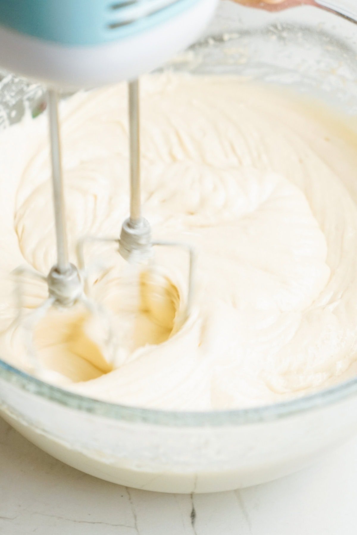 A hand mixer is blending a creamy batter for Christmas cake pops in a clear glass bowl placed on a white countertop.