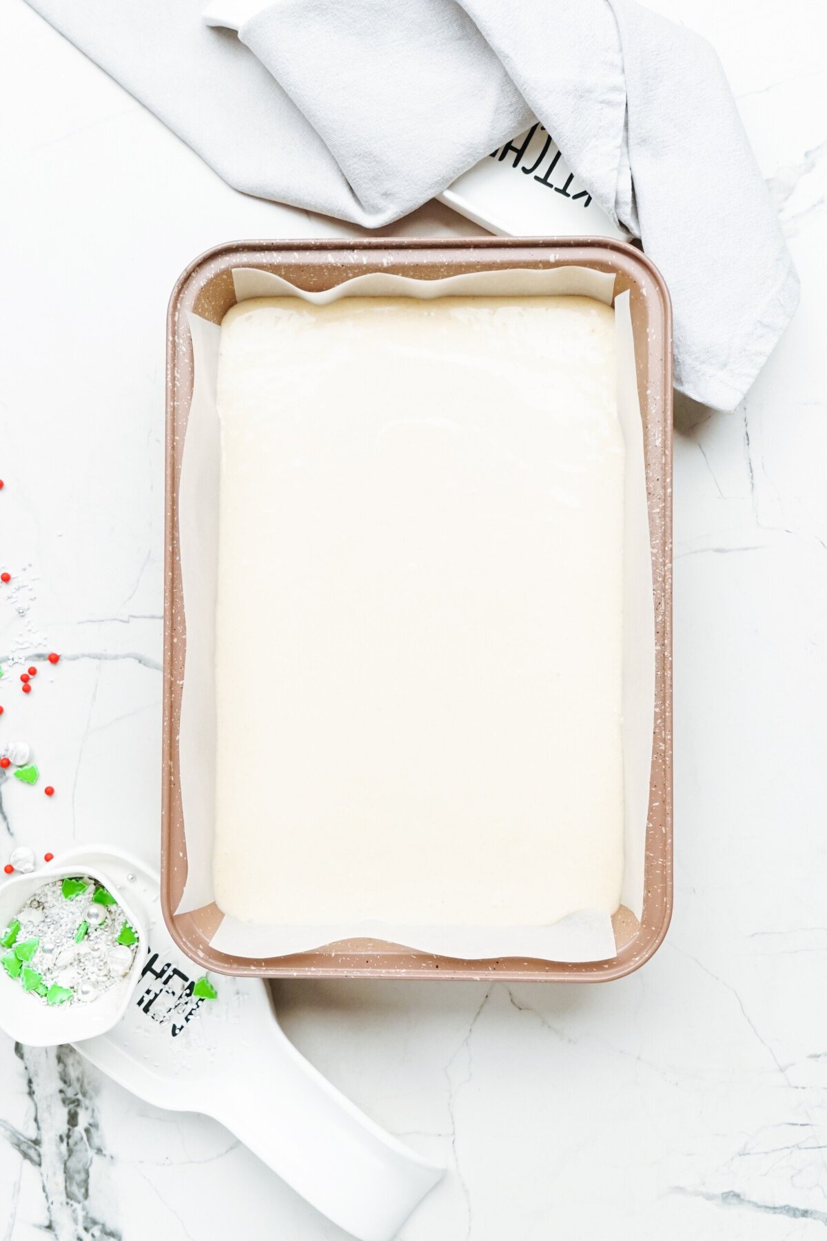 A rectangular baking pan filled with unbaked dough, lined with parchment paper, is placed on a marble countertop. Nearby, a white kitchen cloth and measuring cup with green and red sugar sprinkles await to adorn the festive Christmas cake pops.