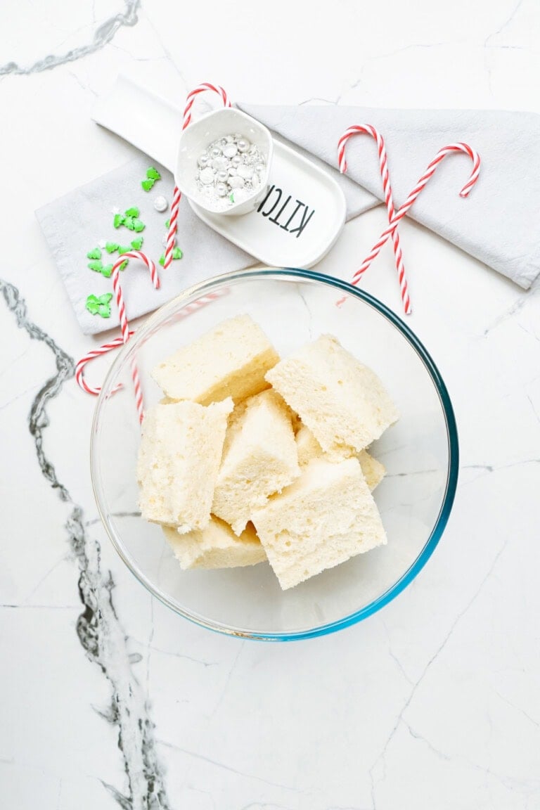 A glass bowl filled with white sponge cake cubes sits on a marble surface, evoking thoughts of delightful Christmas cake pops. Nearby, two candy canes, a small dish of powdered sugar, and a napkin are visible.