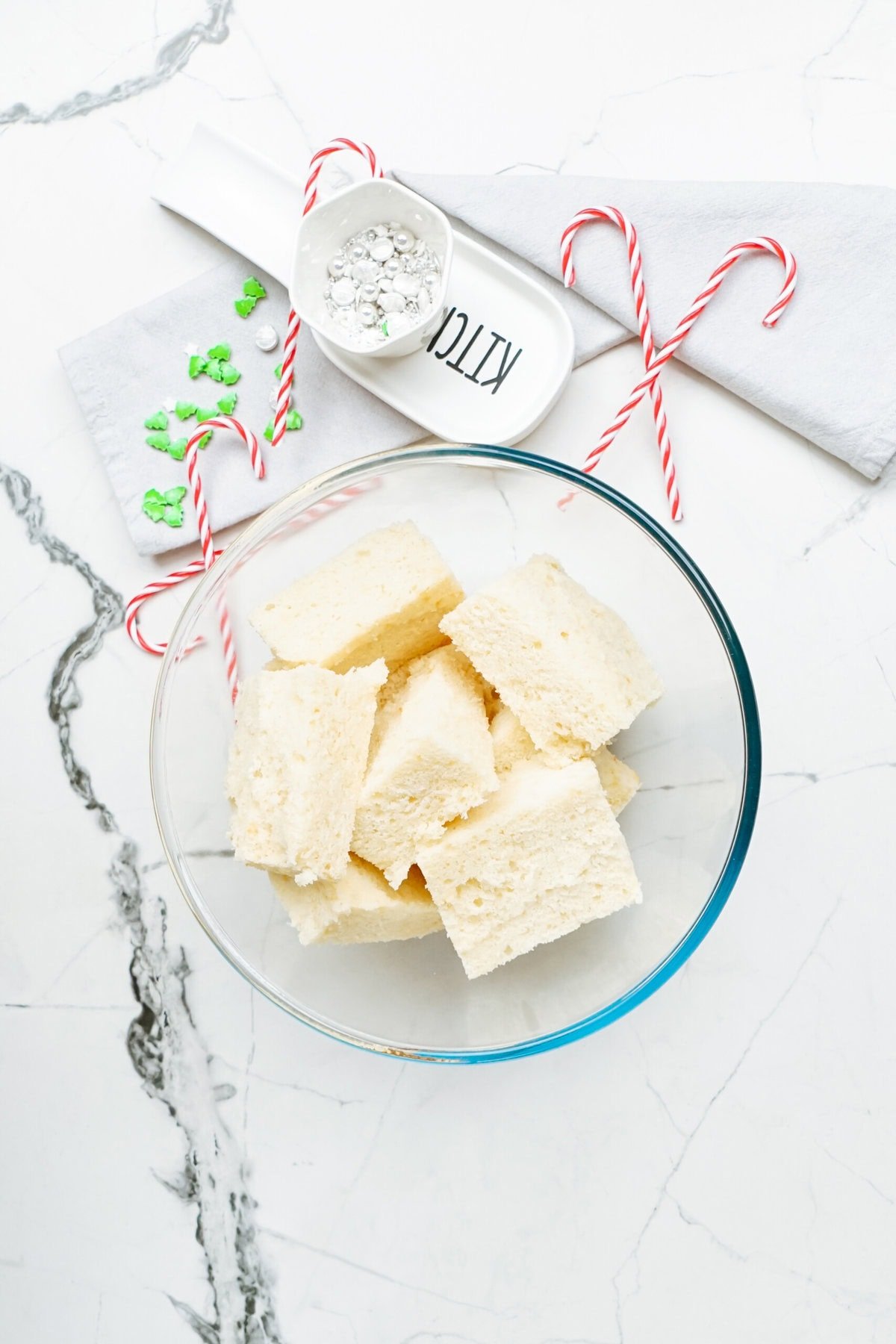 A glass bowl filled with white sponge cake cubes sits on a marble surface, evoking thoughts of delightful Christmas cake pops. Nearby, two candy canes, a small dish of powdered sugar, and a napkin are visible.