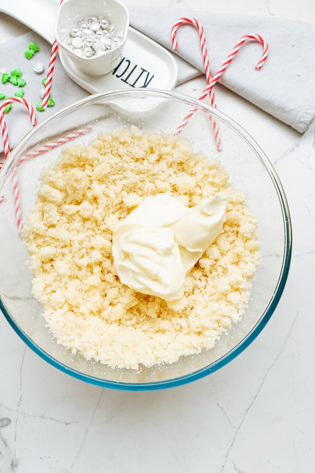 A bowl of crumbly dough with dollops of cream on top, candy canes, and decorating supplies are placed on a marble surface, ready to craft festive Christmas cake pops.