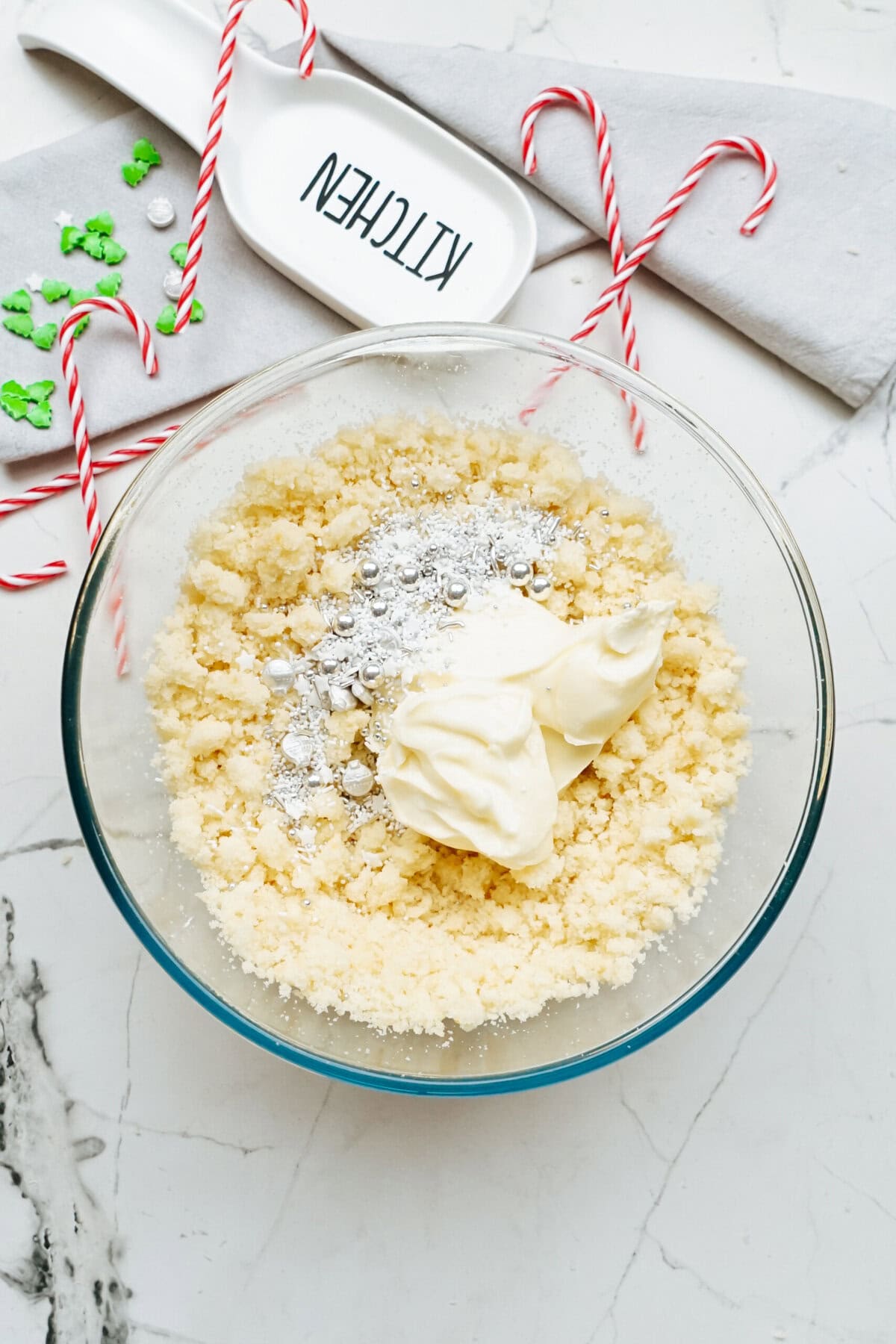 A glass bowl containing cake mix, butter, and cream cheese sits on a marble countertop. Nearby are red and white striped candy canes, hinting at festive Christmas cake pops in the making. A napkin and a spoon with the word "Kitchen" on it complete this cozy holiday scene.