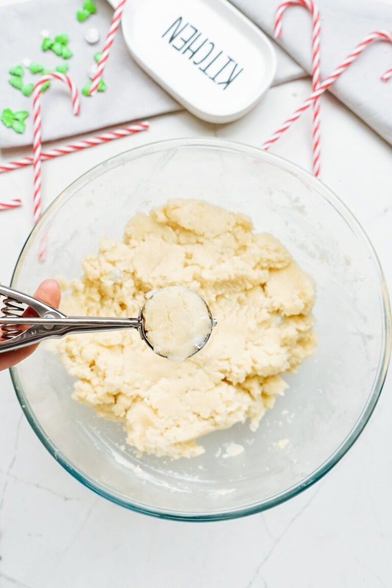 A hand holding a cookie dough scoop over a glass bowl filled with cookie dough. Candy canes and a small dish labeled "KITCHEN" are in the background, ready for festive creations like Christmas cake pops.