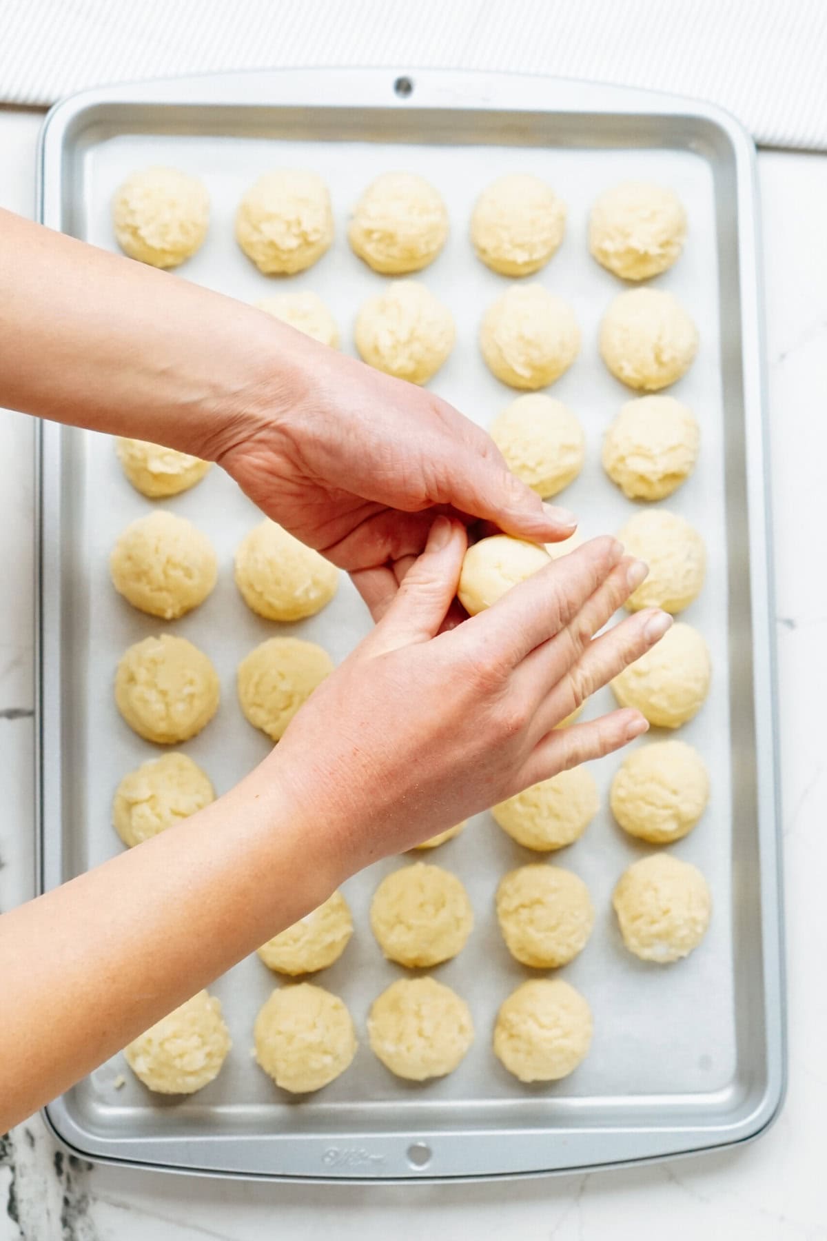 Close-up of hands shaping dough above a baking sheet filled with evenly spaced, unbaked dough balls, destined to become delightful Christmas cake pops.