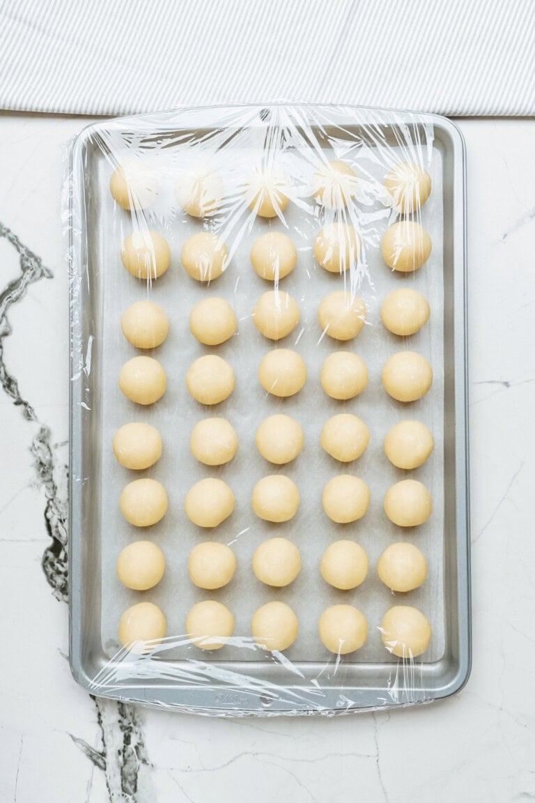 A baking sheet with evenly spaced dough balls, destined to become delightful Christmas cake pops, covered with plastic wrap and placed on a marble countertop.