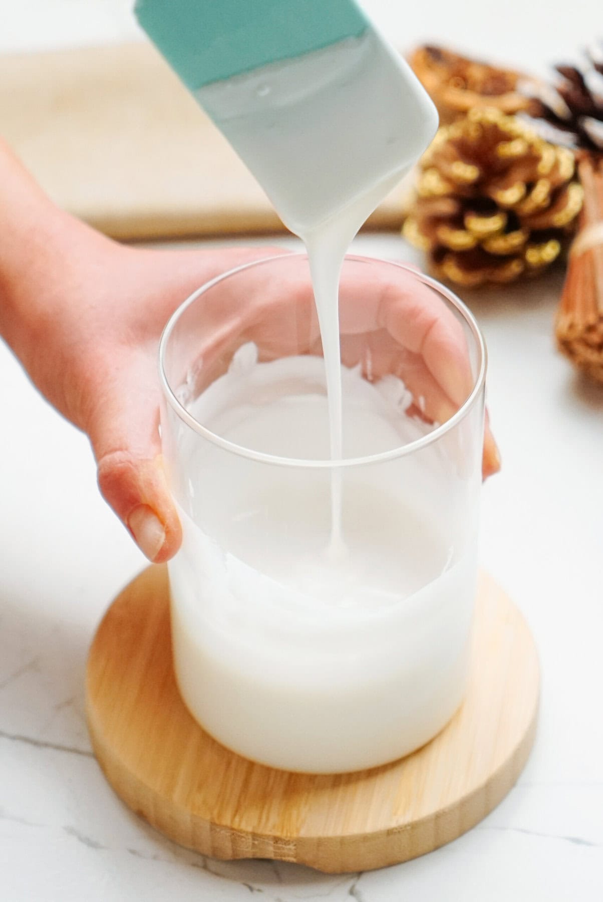 A person pours liquid white soap into a glass container with a wooden coaster, while pinecones, cinnamon sticks, and Christmas cake pops are in the background.