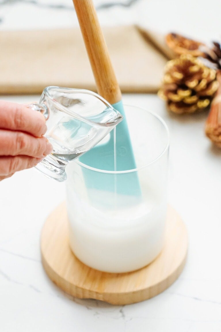 A hand pours liquid from a small pitcher into a glass container with a white substance, likely prep for Christmas cake pops. A spatula is placed inside the container, and pine cones are visible in the background.