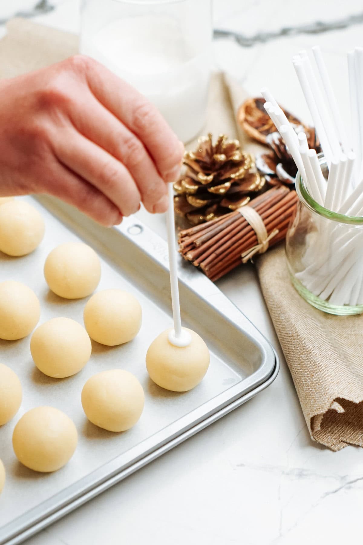 A hand is inserting a stick into dough balls arranged on a baking sheet, preparing festive Christmas cake pops. Nearby are pinecones, a jar of more sticks, and a glass of milk.