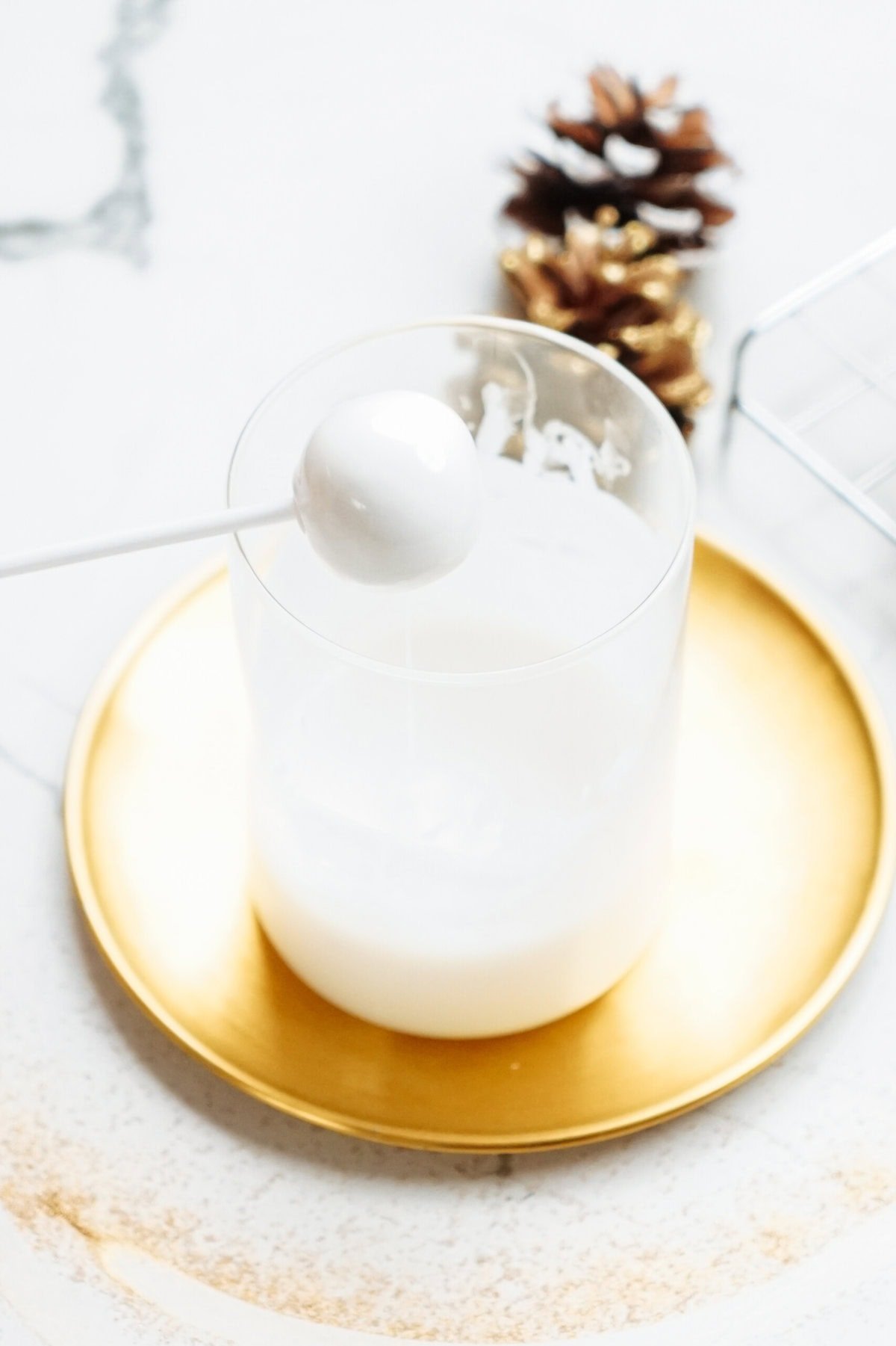 A festive Christmas cake pop on a stick is being dipped into a glass containing white liquid, placed on a gold plate with a pinecone in the background.