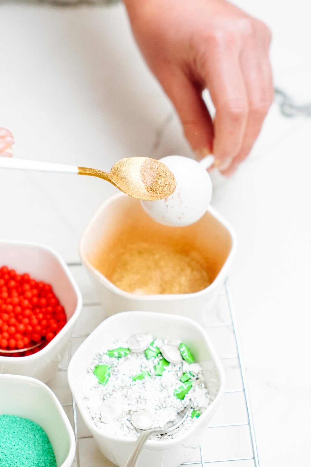 A person decorates a white Christmas cake pop by sprinkling gold powder using a spoon. Nearby bowls contain red sprinkles, green and white candies, and green sugar.