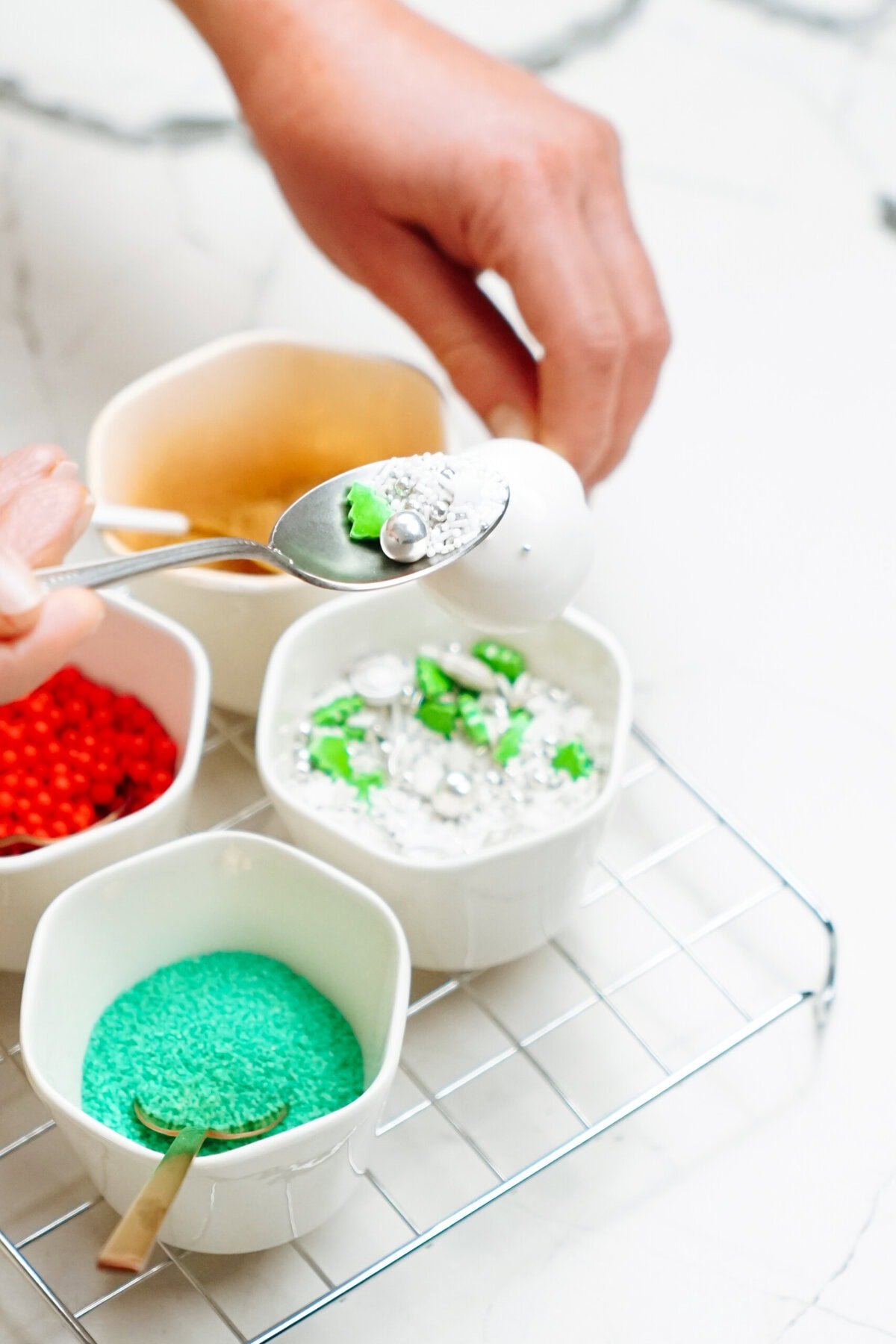 A hand holding a spoon sprinkles green and white decorations onto a dessert, with bowls of various colored sprinkles on a wire rack below. The scene evokes the festive spirit of making Christmas cake pops.