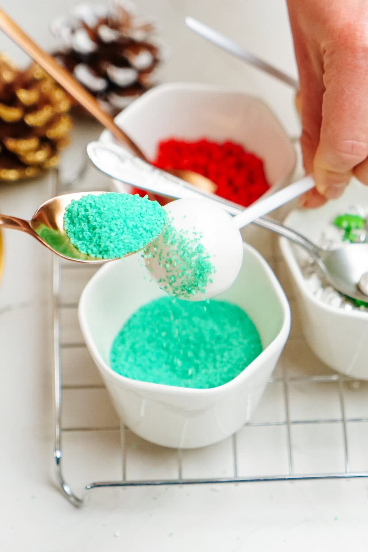 A person sprinkles green sugar onto a white cake pop over a bowl. Nearby are bowls of red sprinkles and other decorations, creating festive Christmas cake pops.