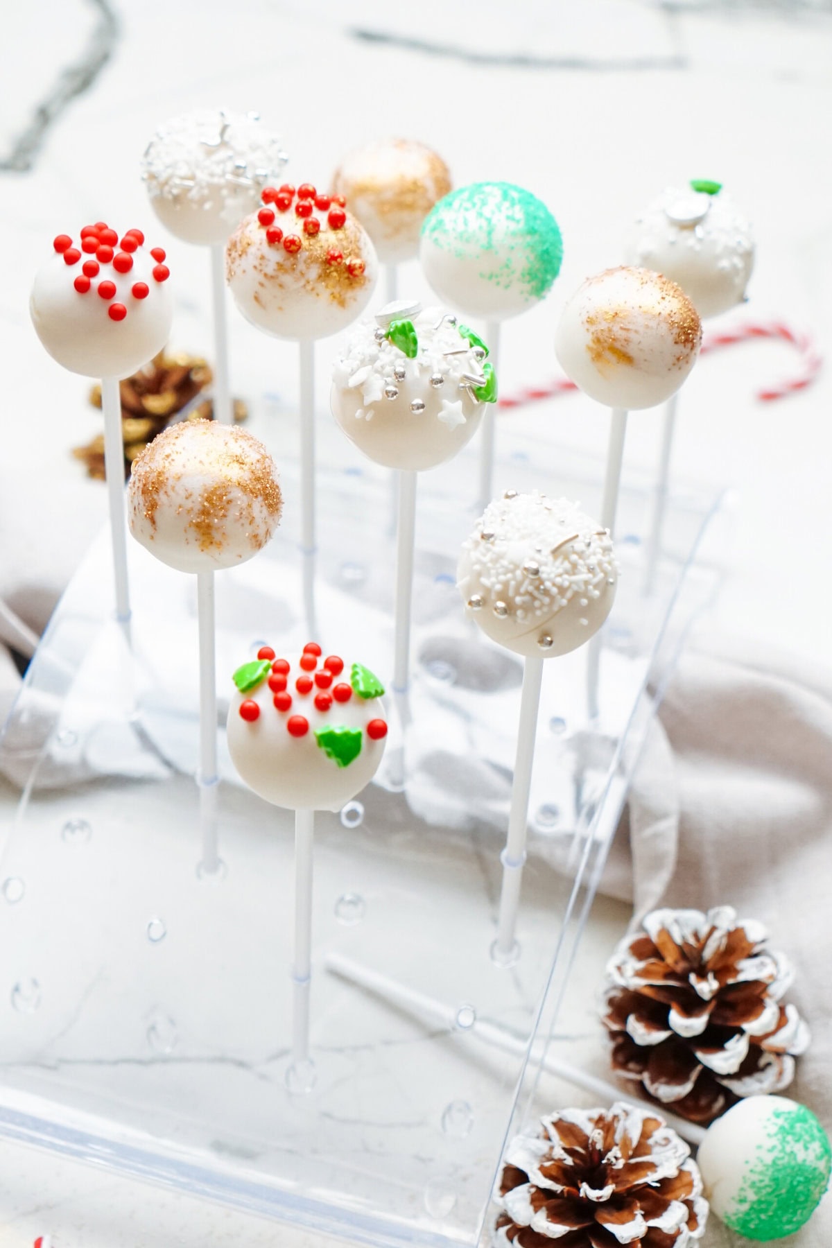 Elegant Christmas cake pops decorated with various sprinkles and coatings, displayed on a transparent stand beside pinecones.