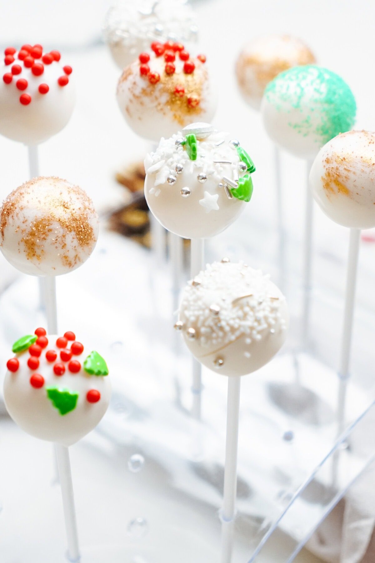 Christmas cake pops decorated with red, green, and silver sprinkles are displayed on sticks in a clear stand.