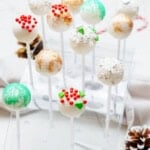 A display of various Christmas cake pops in festive decorations, including red, green, and white sprinkles, set in a clear stand on a white surface with pinecones.