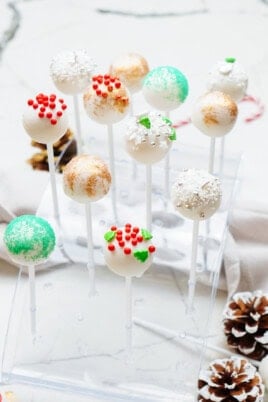 A display of various Christmas cake pops in festive decorations, including red, green, and white sprinkles, set in a clear stand on a white surface with pinecones.