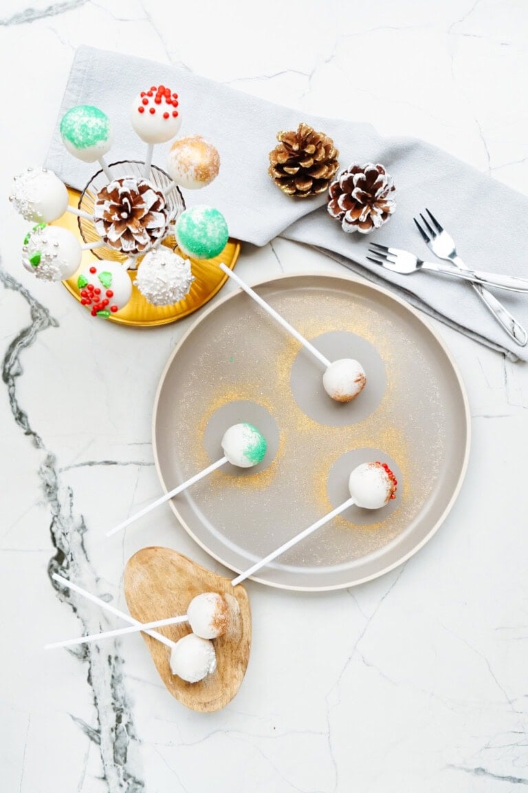 A display of assorted Christmas cake pops on a marble surface, with some on a beige plate and others on a yellow tray. Pinecones and forks are also part of the festive decor.