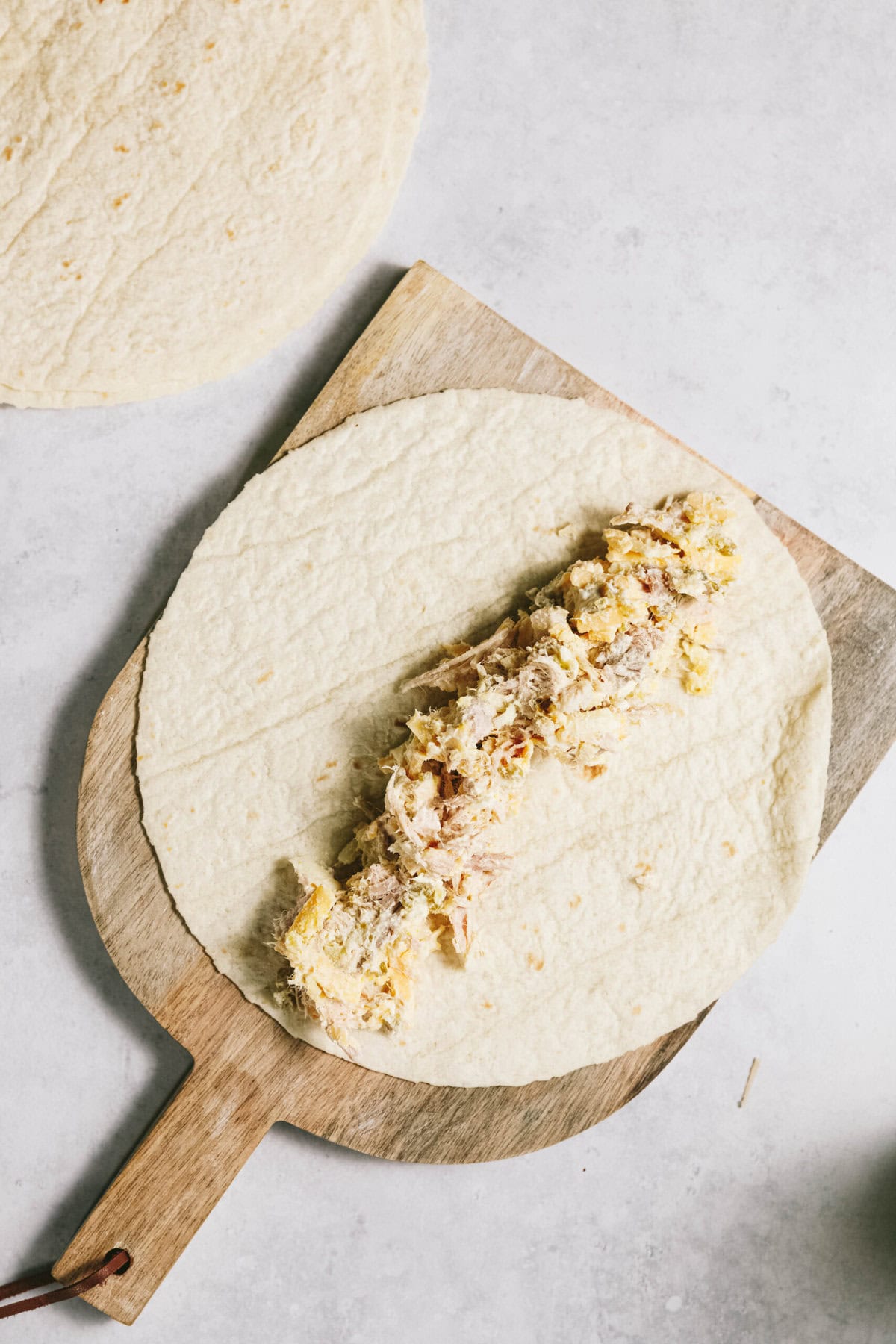 A flour tortilla with a filling, placed on a wooden board. An additional tortilla is partially visible in the background.