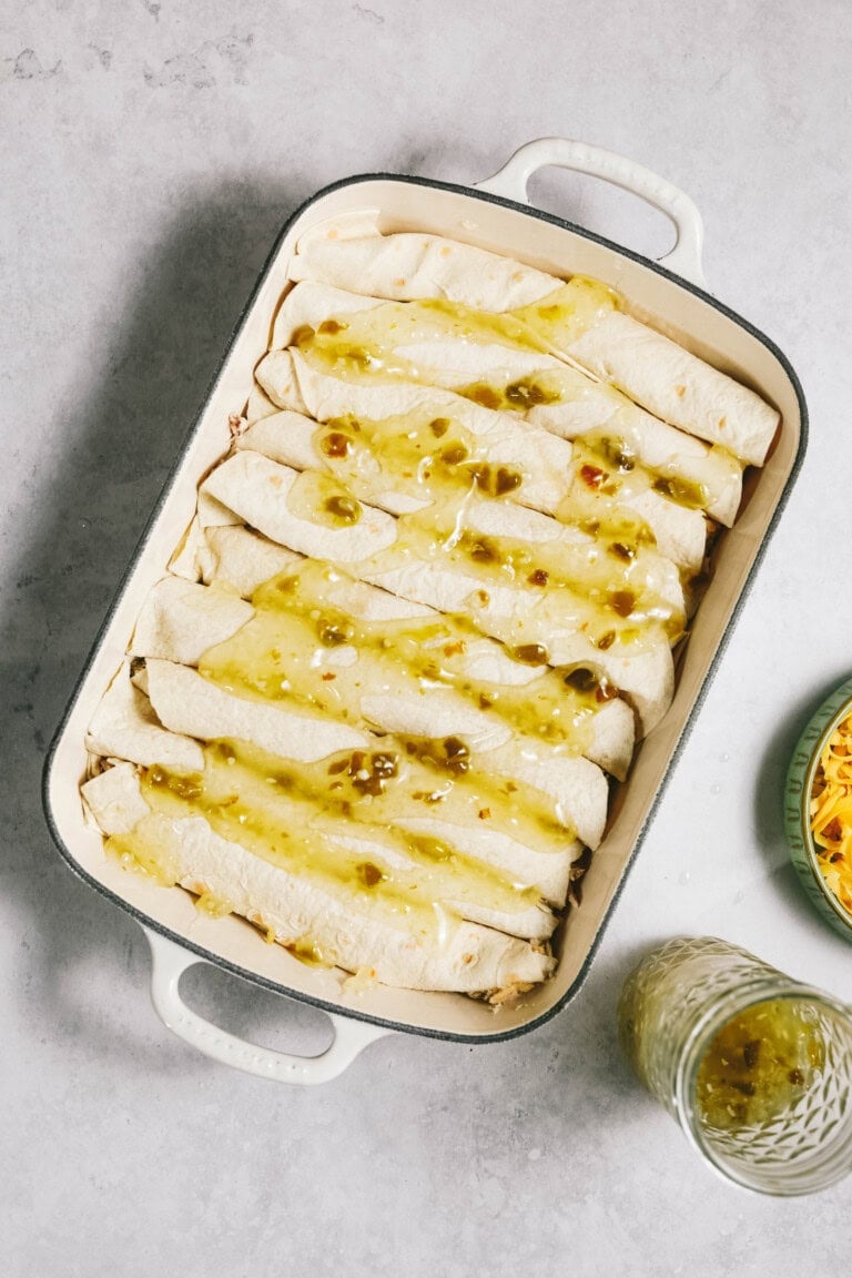 A rectangular baking dish filled with rolled enchiladas topped with green sauce, placed on a gray surface next to a jar of more green sauce and a bowl of shredded cheese.