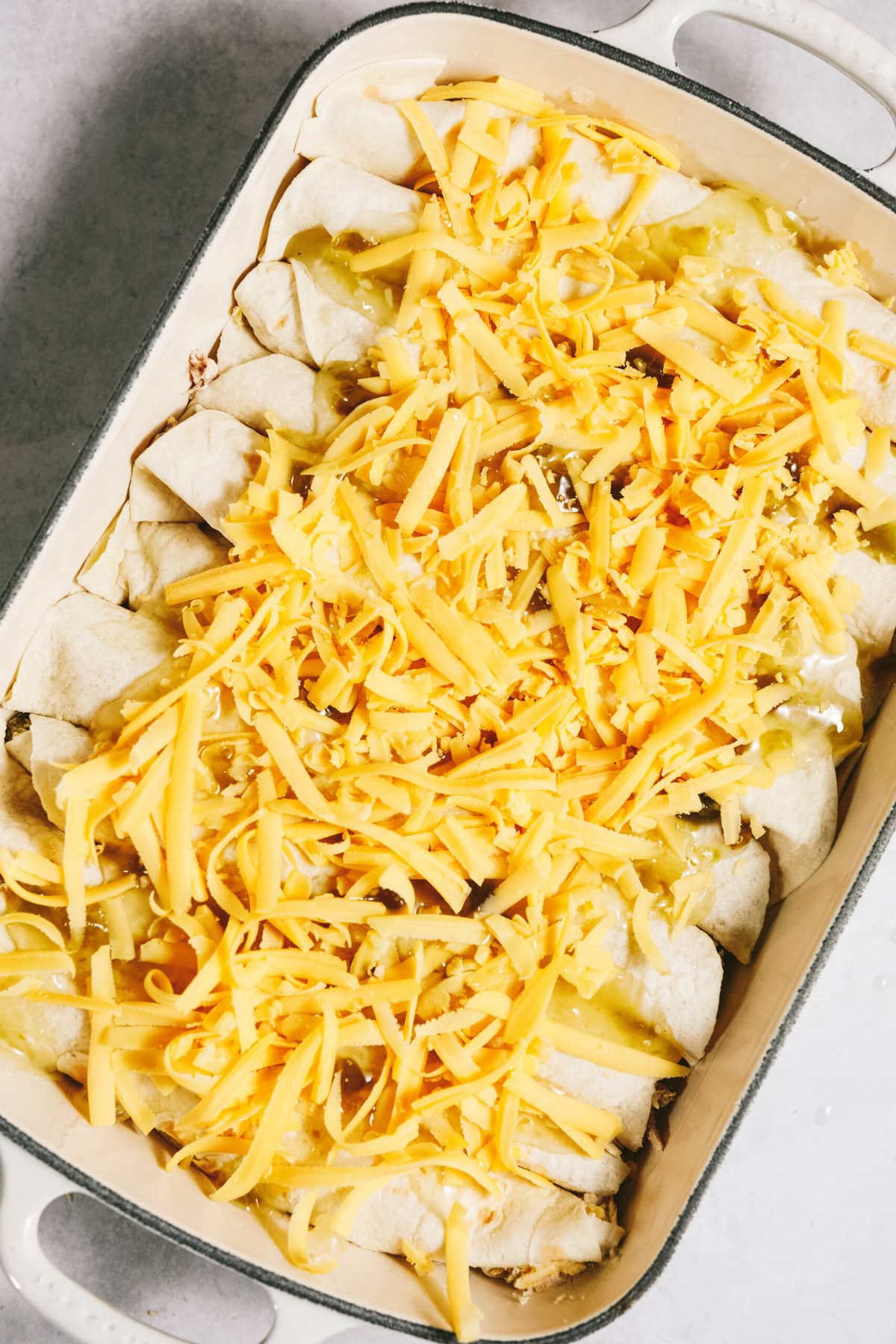 A baking dish filled with rolled tortillas topped with grated cheese and sauce, ready to bake.