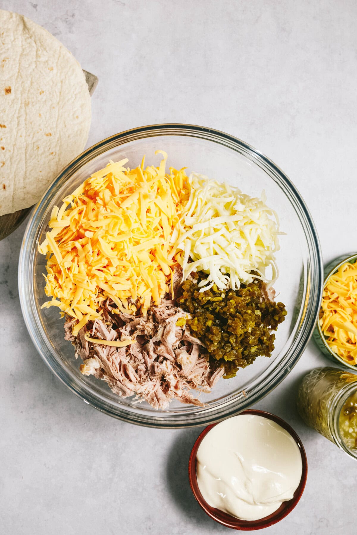 A container containing grated cheeses, shredded meat, and chopped vegetables, placed on a light surface next to a tortilla, salsa, and a small bowl of sour cream.