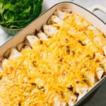 A baking dish filled with enchiladas topped with melted cheese, with a bowl of fresh cilantro leaves and a jar of sauce in the background.