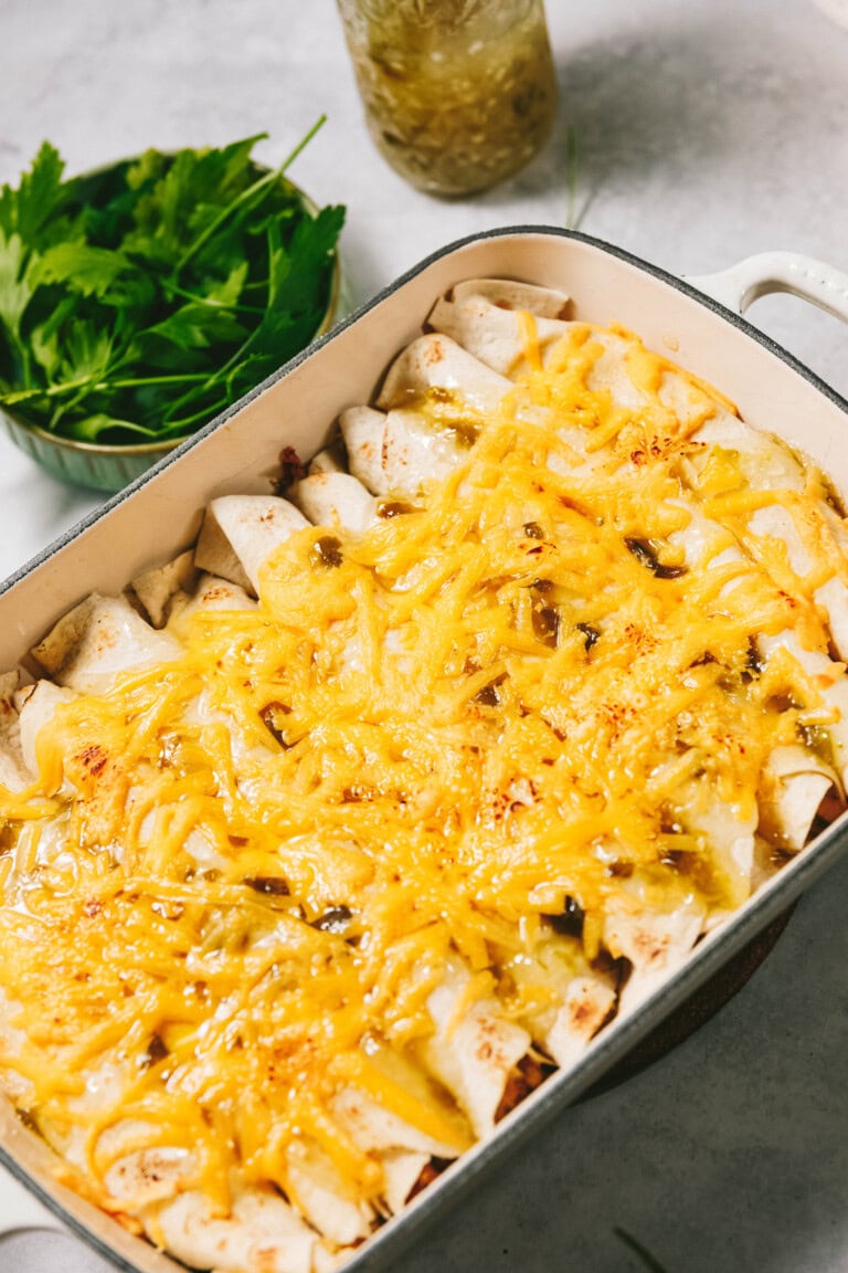 A baking dish filled with enchiladas topped with melted cheese, with a bowl of fresh cilantro leaves and a jar of sauce in the background.